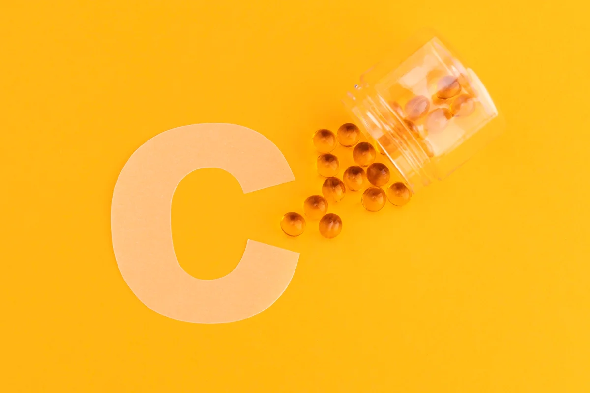 Foto colorida com fundo laranja, a imagem da letra C e um pote transparente com cápsulas redondas de vitamina C - Metrópoles.