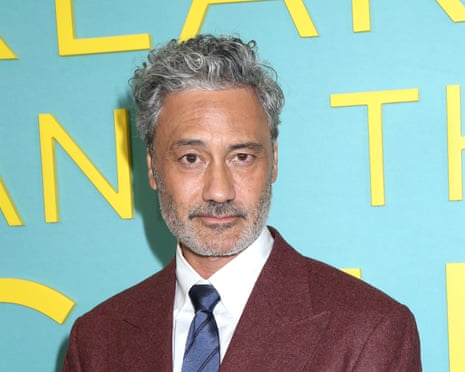 Taika Waititi, with grey curly hair and a beard, wears a maroon blazer and striped tie