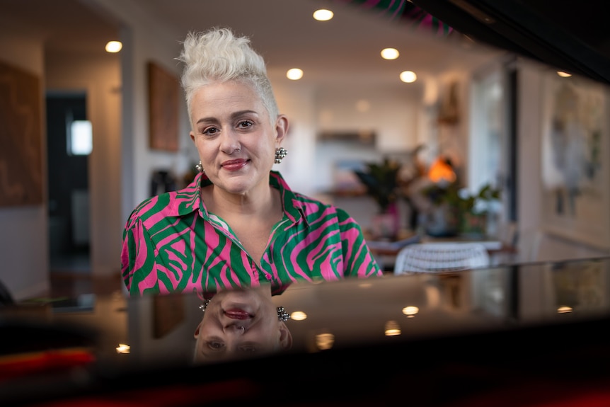 Katie Noonan with short blonde hair wears a colourful shirt sitting behind a piano that has a reflective surface.
